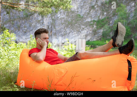 Freelance work or study concept. young man in casual wear using laptop and talking on smartphone. sitting in an inflatable chair on the grass with a l Stock Photo