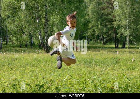 9 years old boy in uniform plays football on a green meadow. A child kicks a feint with a soccer ball, throws it Stock Photo