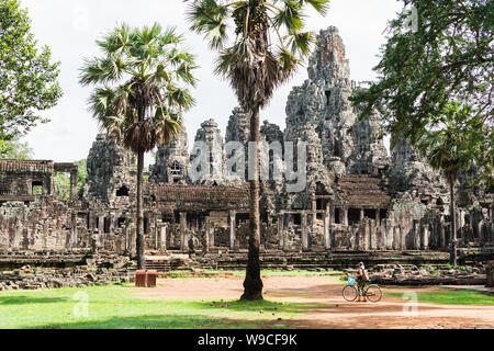 Young woman riding bicycle next to ancient Bayon temple ruins in Angkor Wat complex, Cambodia Stock Photo