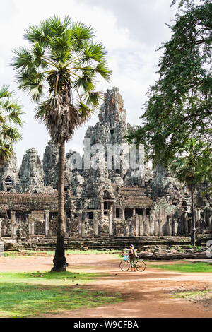 Young woman riding bicycle next to ancient Bayon temple ruins in Angkor Wat complex, Cambodia. Vertical orientation Stock Photo