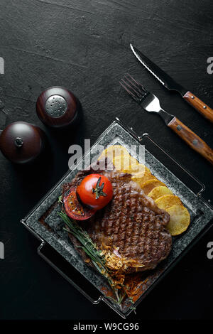 Grilled steak with knife and fork carved on black stone slate. Steak on a hot marble stone. Copy space, dark background, food fashion photo. Stock Photo