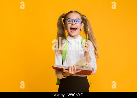 Excited Elementary Schoolgirl Holding Book And Pencil On Yellow Background Stock Photo