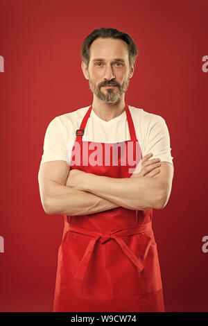 Confident in his culinary craft. Cook with beard and mustache wearing apron red background. Man mature cook posing cooking apron. Chief cook and professional culinary. Cook food at home. Fine recipe. Stock Photo