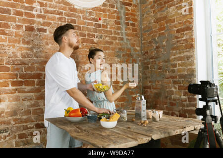 Vlogger and blogger freelance job, food concept. Young caucasian couple cooking together and recording live video for vlog and social media with professional camera against brick wall in their kitchen. Stock Photo