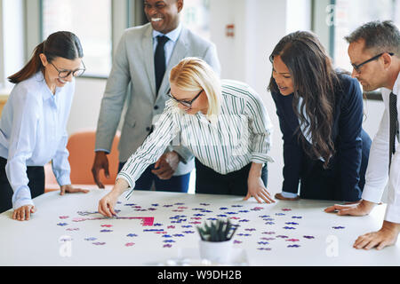 Smiling young businesswoman putting puzzle pieces together on an office table surrounded by a diverse group of laughing coworkers Stock Photo