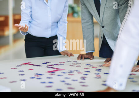 Closeup of a diverse group of smiling businesspeople standing together around a boardroom table in an office trying to solve a jigsaw puzzle Stock Photo