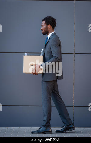 side view of fired african american businessman holding carton box while walking on street Stock Photo