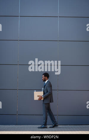 side view of fired african american businessman carrying carton box while walking along wall Stock Photo