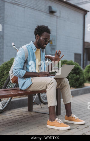 cheerful african american man waving hand while sitting on bench during video call in laptop Stock Photo