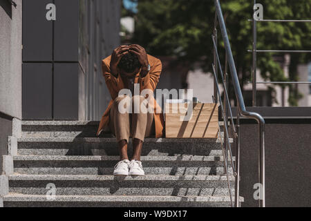 depressed african american businessman sitting on stairs near cardboard box and holding hands on head Stock Photo