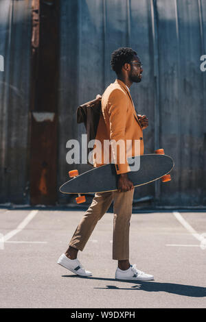 stylish african american businessman holding longboard while walking on rooftop Stock Photo