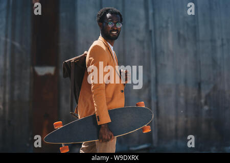 stylish african american businessman holding longboard and smiling at camera Stock Photo