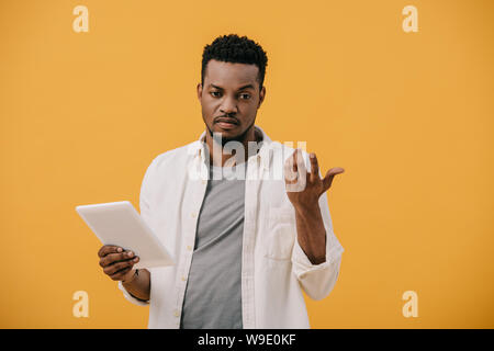 upset african american man gesturing while holding digital tablet isolated on orange Stock Photo