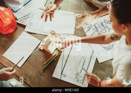 Top view of achitect-engineer's hands shows future house, office or store design plans to a young couple. Meeting at the construction office to talk about facade, interior decoration, home layout. Stock Photo