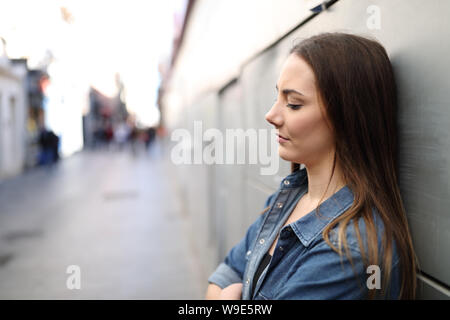 Side view portrait of a sad girl alone complaining leaning on a wall in a solitary street Stock Photo