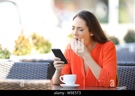 Tired woman yawning holding smart phone in a bar terrace Stock Photo