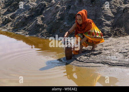 girl of oriental appearance in sari and hijab fills the pitcher with water from a dirty source in the desert Stock Photo