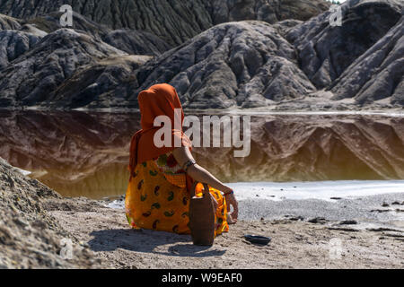 young woman in ethnic attire sits with a jug on the shore of a lake in a deserted valley Stock Photo