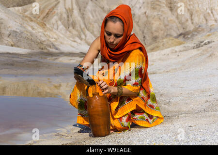 girl of oriental appearance in sari and hijab fills the pitcher with water from a dirty source in the arid area Stock Photo