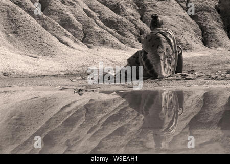 young woman in ethnic attire sits with a jug on the shore of a lake in a deserted valley Stock Photo