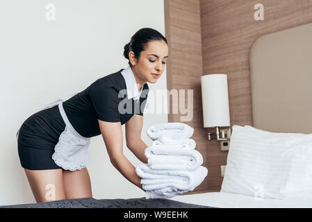 pretty maid in white apron putting pile of folded towels on bed in hotel room Stock Photo