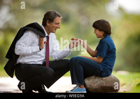 Businessman father crouches down to knock fists together (daps) with his young son who sits on a rock in a park. Stock Photo