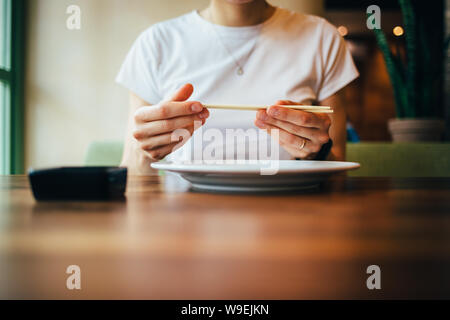 Close-up of female hands holding chopsticks. Young woman is preparing to eat sushi sitting at table in restaurant. Stock Photo