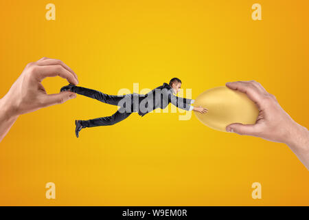 Small businessman being pulled in opposite directions by two big hands, and clutching with his both hands at gold egg in one of the hands. Stock Photo