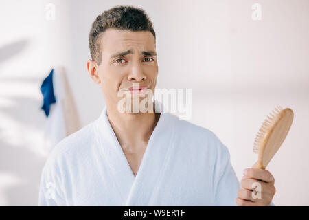 disappointed young man in bathrobe holding hairbrush, looking at camera and making grimace Stock Photo