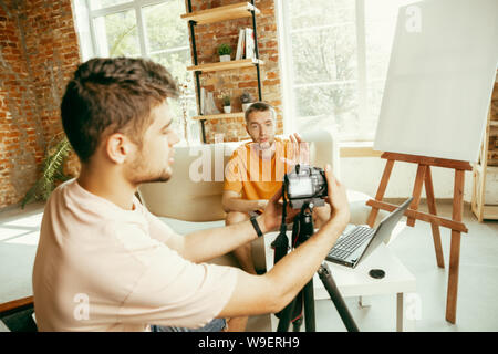 Two young caucasian male bloggers in casual clothes with professional equipment or camera recording video interview at home. Blogging, videoblog, vlogging. Talking while streaming live indoors. Stock Photo