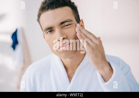 serious young man in bathrobe touching face and looking at camera Stock Photo