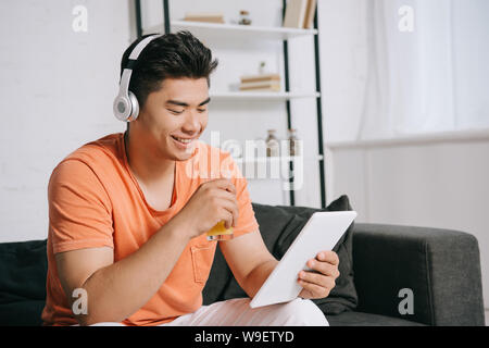cheerful asian man drinking orange juice while using digital tablet and listening music in headphones Stock Photo