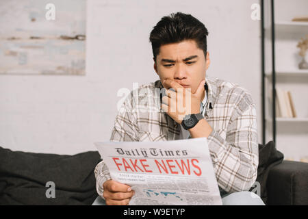 serious asian man reading fake news newspaper while sitting on sofa at home Stock Photo