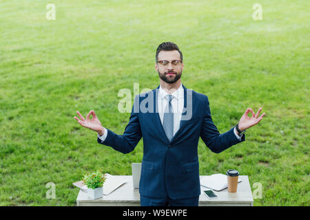 young businessman in formal wear meditating near table with various office stuff in park Stock Photo