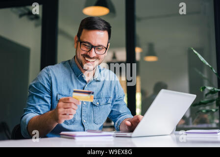 Happy cheerful smiling young adult man doing online shopping or e-shopping satisfied entrepreneur making online payment paying for service or goods se Stock Photo