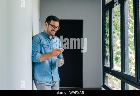 Confident smiling young cheerful casual man looking in and using his mobile smart phone satisfied with content drinking coffee leaning on wall near bi Stock Photo