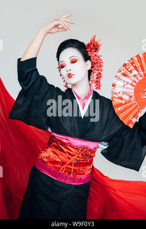 beautiful geisha in black kimono with hand fan dancing near red cloth on background isolated on white Stock Photo