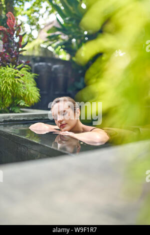 Healthy woman relaxing outdoors in sunny and plant surrounded swimming pool of luxurious hotel during tropical vacation in Bali Stock Photo