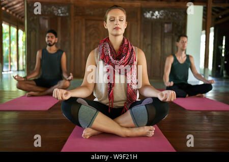 Beautiful woman teaching meditation to two multi-ethnic men in lotus pose on yoga mats in traditional temple in Bali Indonesia