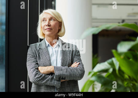 Mature female leader in formal clothing standing with arms crossed and looking through the window she thinking about something Stock Photo