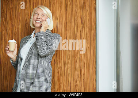 Mature beautiful businesswoman in jacket talking on mobile phone and laughing while drinking coffee from disposable cup at office Stock Photo