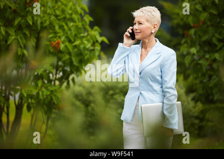 Mature elegant businesswoman holding laptop computer and listening to somebody on mobile phone while walking in the park Stock Photo