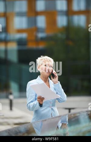 Mature beautiful businesswoman holding documents and talking on mobile phone while crossing the road in the city Stock Photo