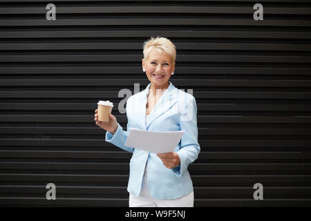 Portrait of successful businesswoman holding documents and coffee drink and smiling at camera outdoors Stock Photo
