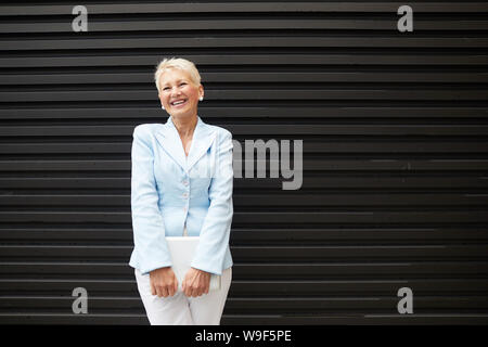Portrait of mature successful businesswoman with laptop standing outdoors in the city and laughing Stock Photo