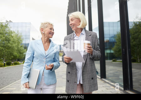 Two successful mature businesswomen discussing new project while going along the street and drinking coffee Stock Photo