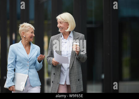 Two mature elegant businesswomen holding documents discussing business contract and laughing while walking in the city Stock Photo