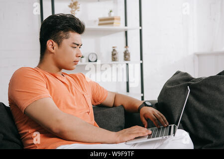 young, attentive asian man using laptop while sitting on couch at home Stock Photo