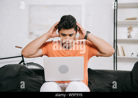 shocked asian man holding hands near head while sitting on sofa and using laptop Stock Photo