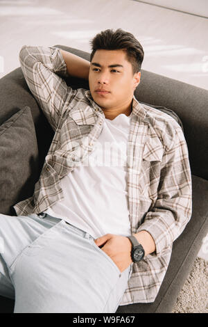 young, dreamy asian man looking away while lying on sofa at home Stock Photo